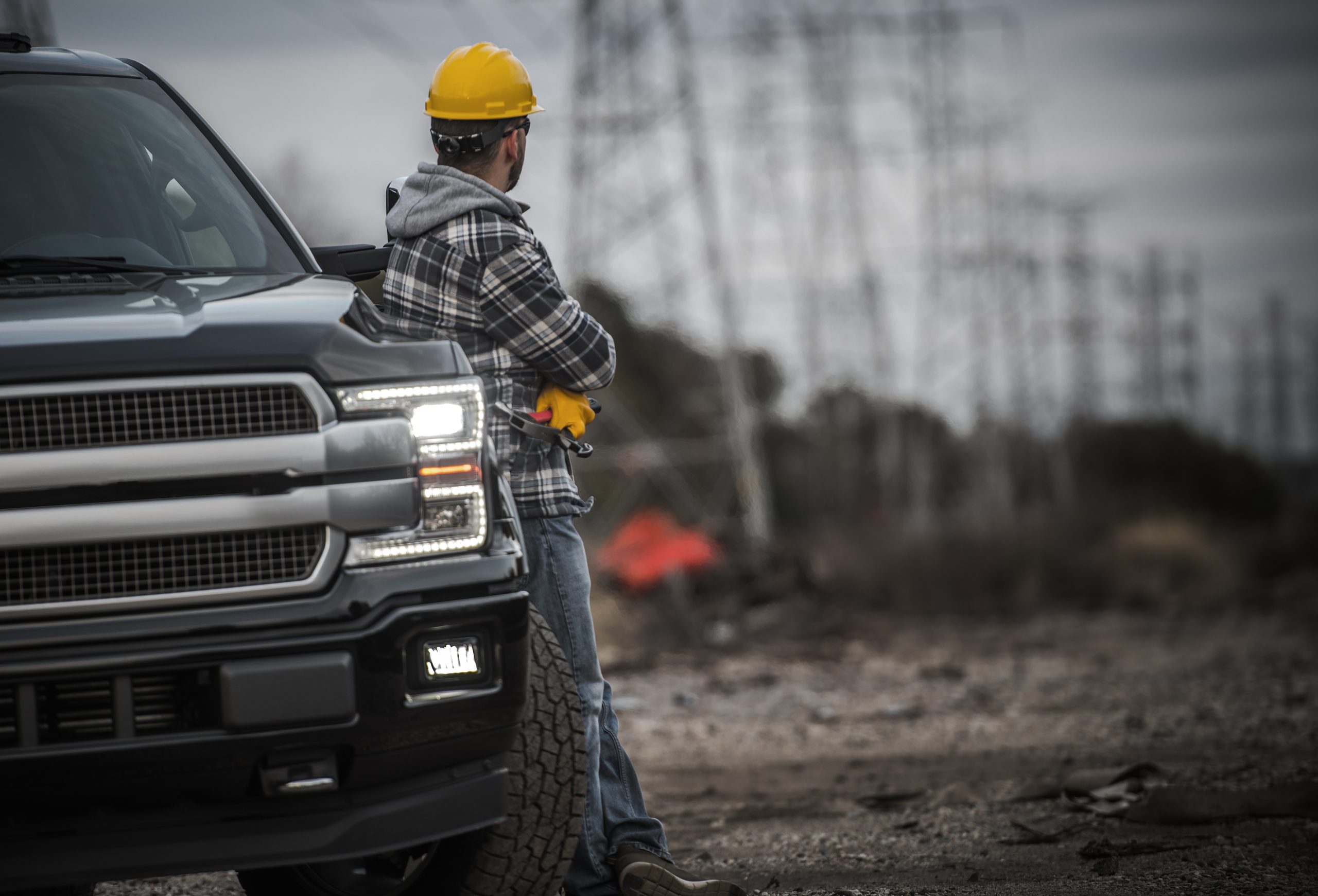 Caucasian Contractor Field Work Job. Worker and His Pickup Truck. Countryside Remote Location with High Voltage Poles in the Background.